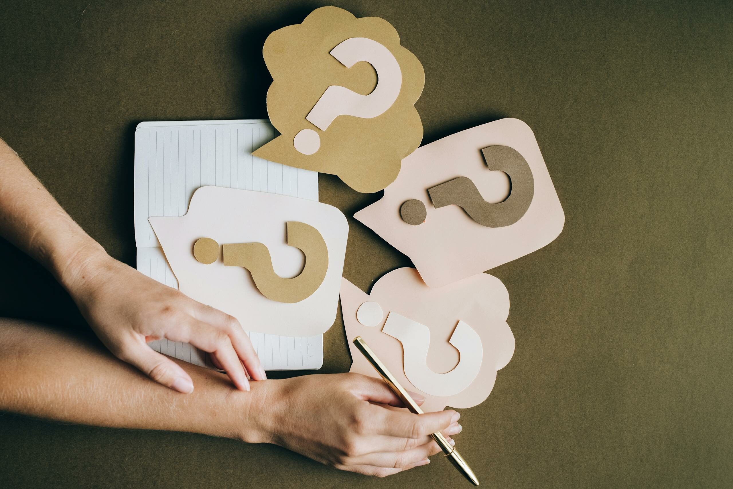 Hands holding pen next to paper question marks on notebook, symbolizing inquiry and creativity.